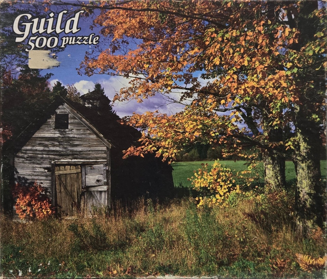 A rustic wooden barn sits in a sunlit meadow during autumn, framed by vibrant orange and green trees.
