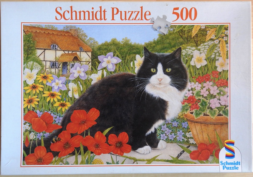 A black and white cat sitting in a vibrant cottage garden filled with flowers and a thatched-roof house in the background.