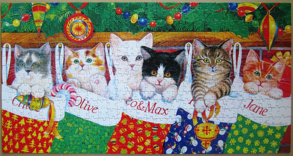Six kittens inside individual Christmas stockings hanging on a mantle with ornaments and greenery above.