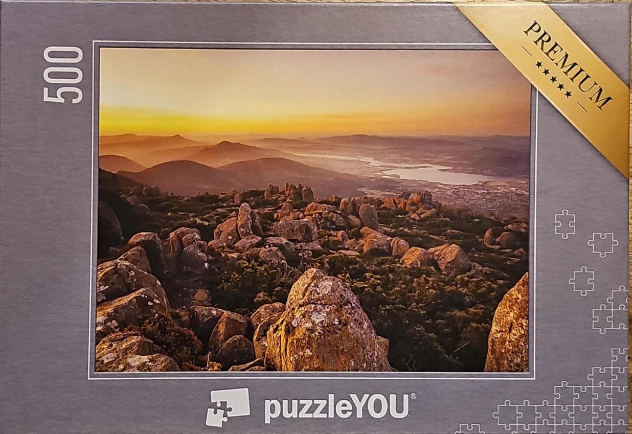 A panoramic landscape photograph of a rocky mountain summit at sunrise overlooking a distant valley and lake.