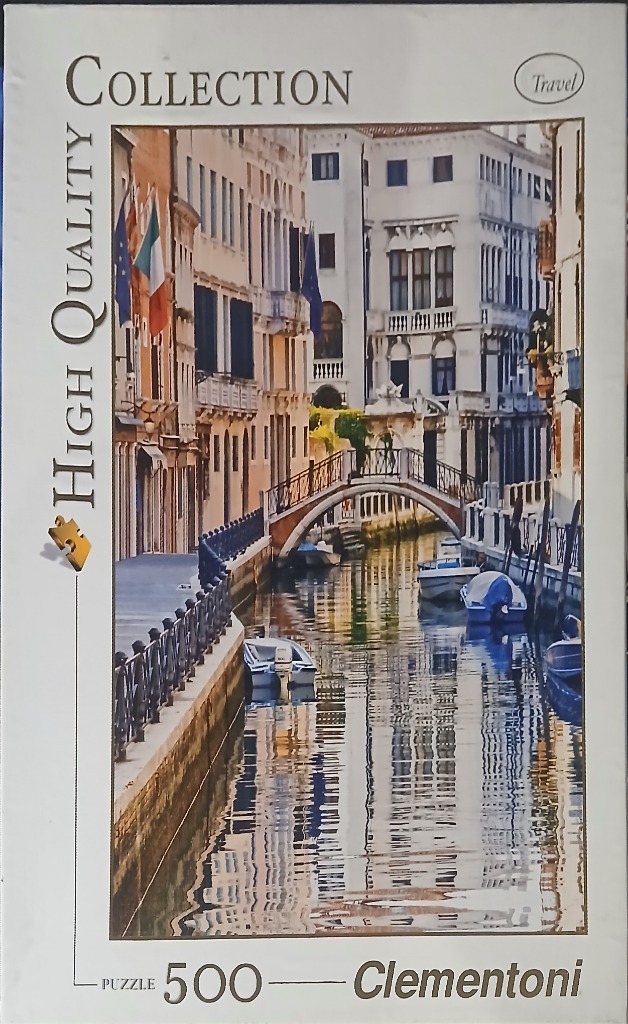 A canal scene in Venice featuring historic buildings, a small arched bridge, and moored boats reflected in the water.
