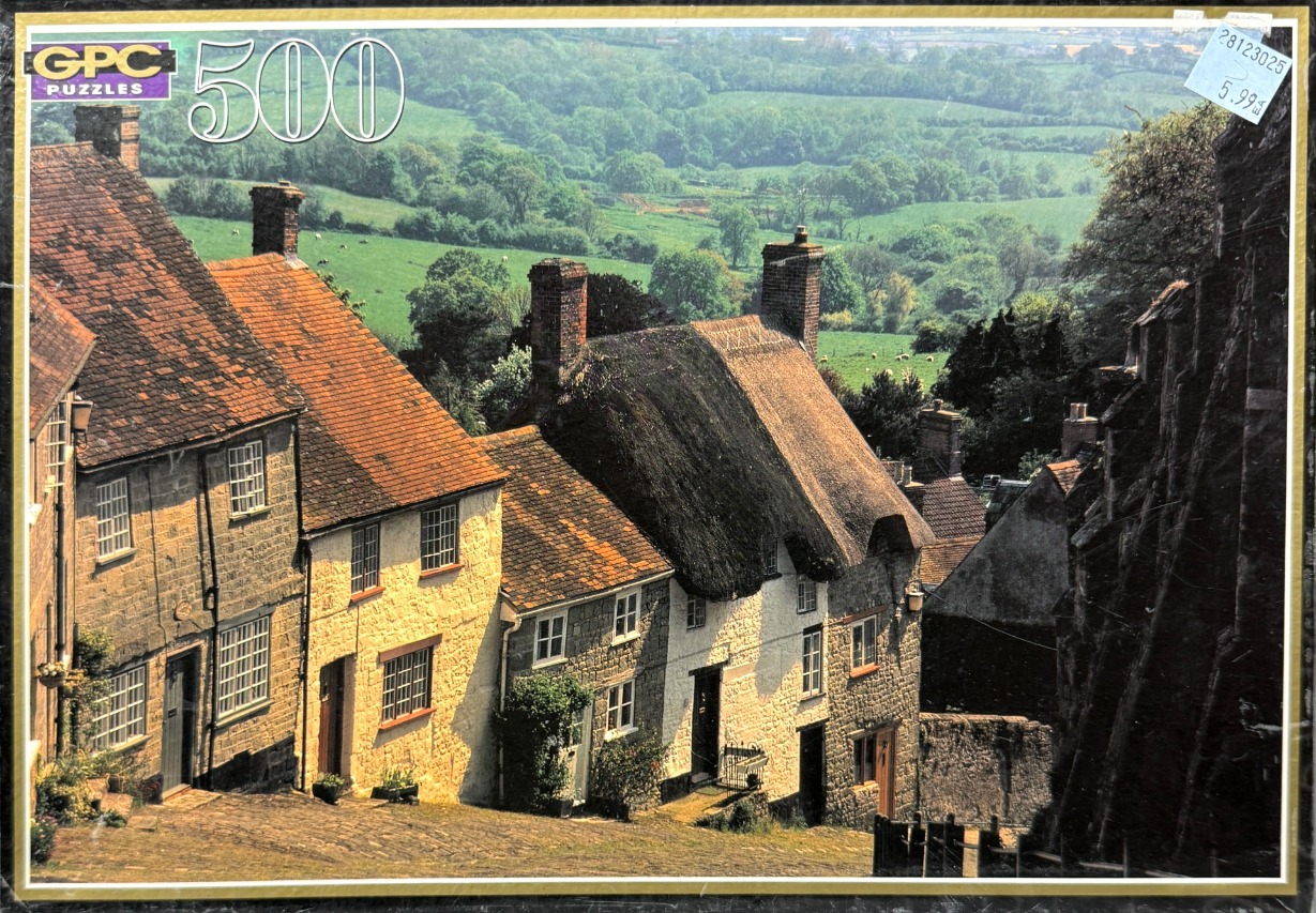 A sunny, picturesque street of stone cottages in the English countryside with a rolling green hill in the background.