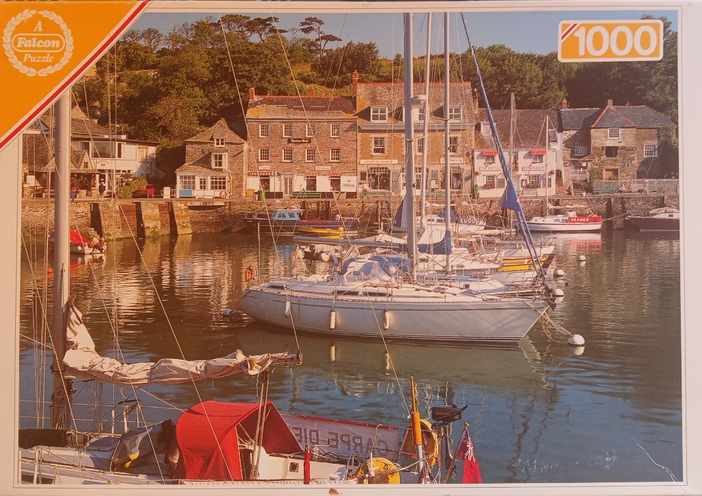 A sunny seaside harbour with sailing boats moored in front of stone buildings and lush green hillside.