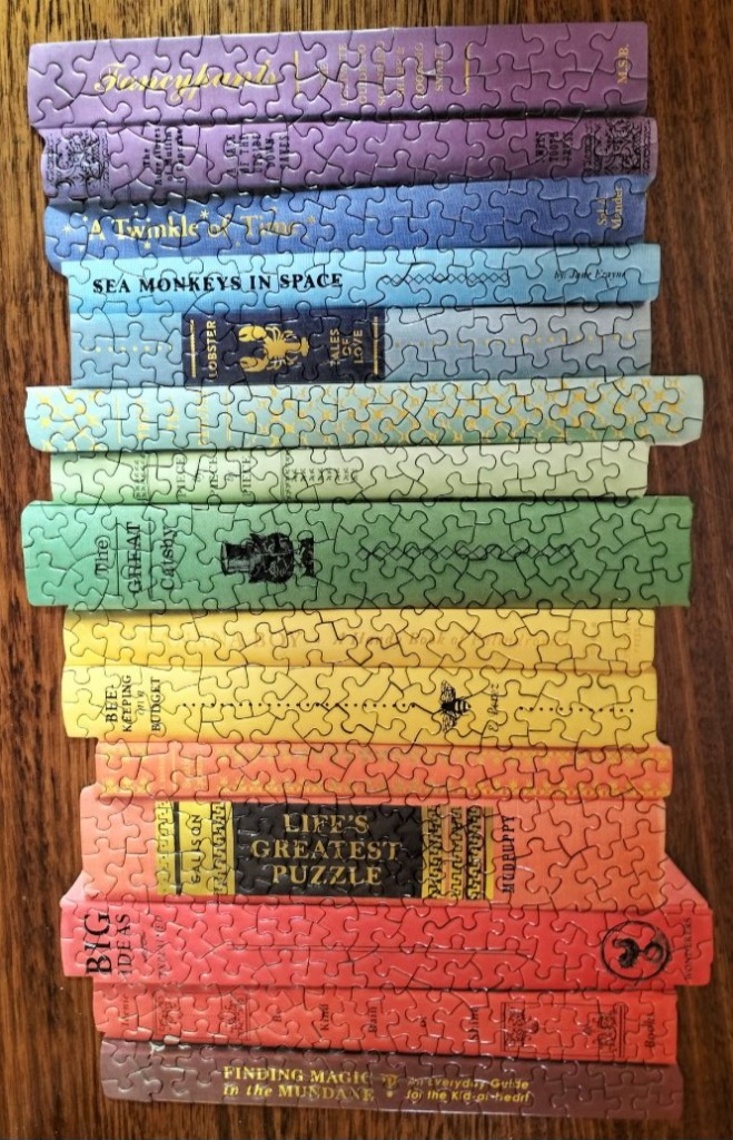 A vertical stack of colorful, whimsical book spines in a rainbow gradient on a wooden surface.