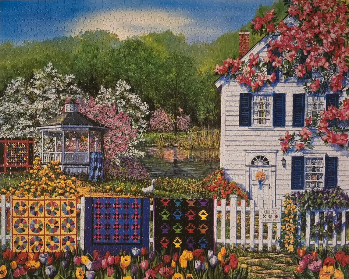 A white house and gazebo in a lush garden with colorful quilts hanging on a picket fence.