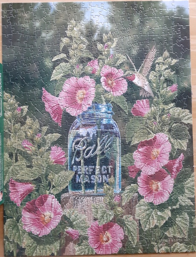 A hummingbird approaches pink hollyhocks in a glass mason jar on a wooden post in a garden.