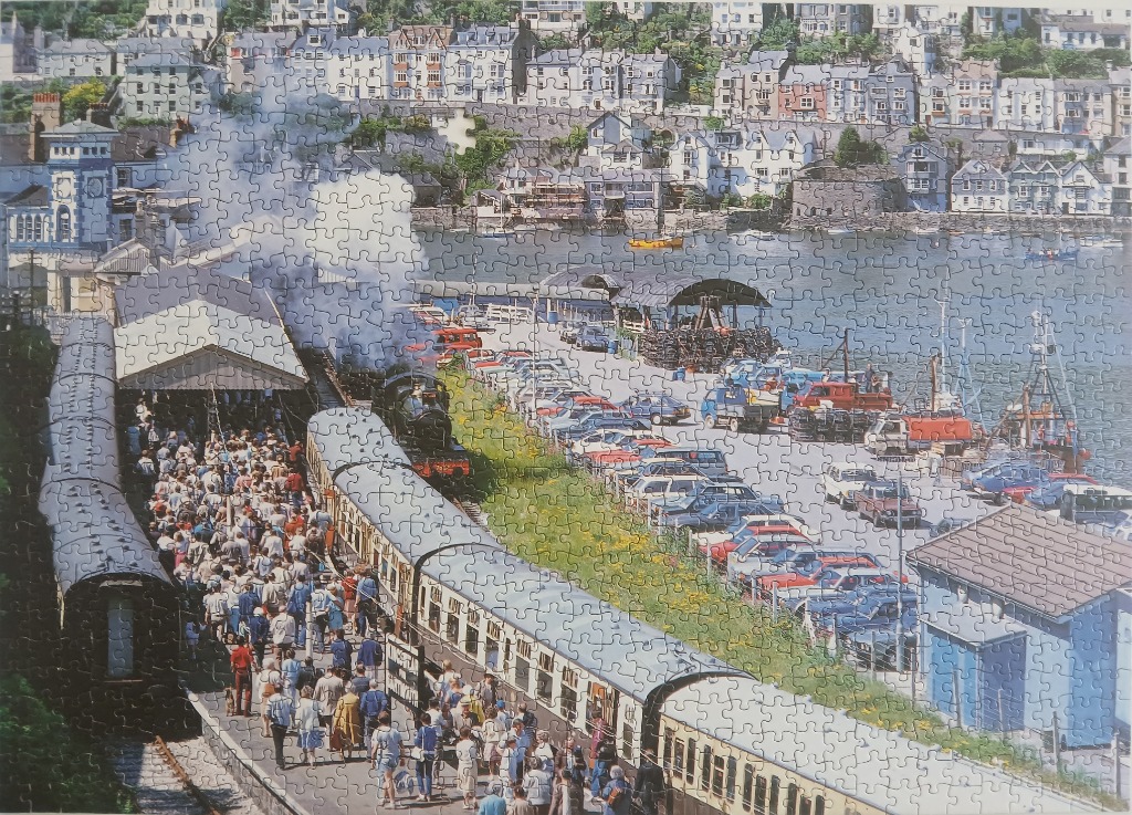 A steam train pulling into a crowded station by a harbour with a hillside town in the background.