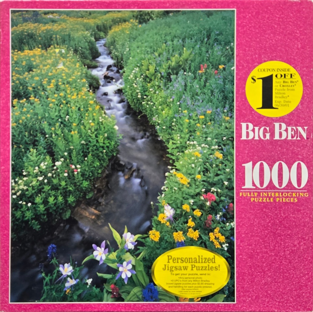 A narrow stream flowing through a lush mountain meadow filled with yellow and purple wildflowers.