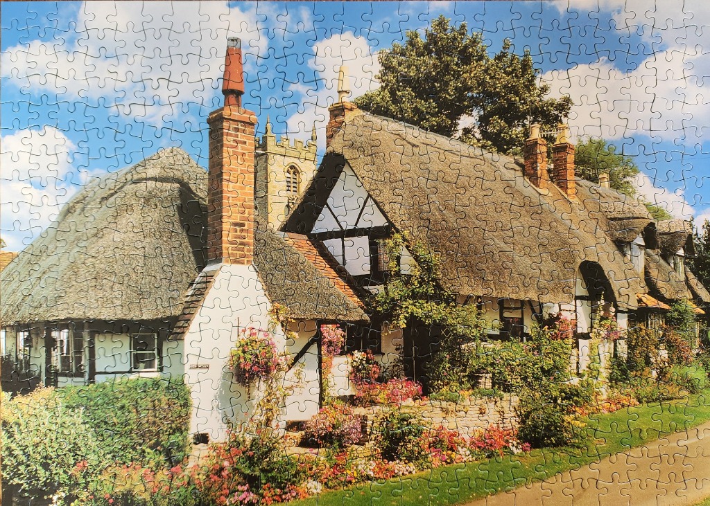 A charming English cottage with a thatched roof and flower-filled garden next to a church tower.