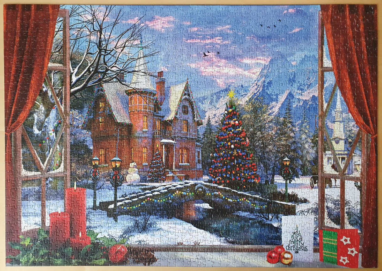 View from a window of a snow-covered village square at Christmas with a Victorian house, tree, and stone bridge.