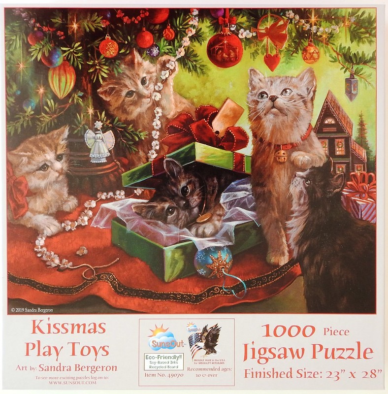 Four kittens playing under a decorated Christmas tree with a green gift box on a red tree skirt.