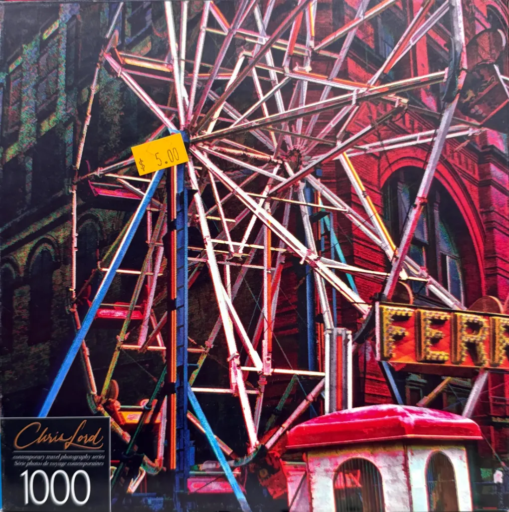 A close-up photograph of a brightly lit Ferris wheel frame against a dark, ornate red brick building.