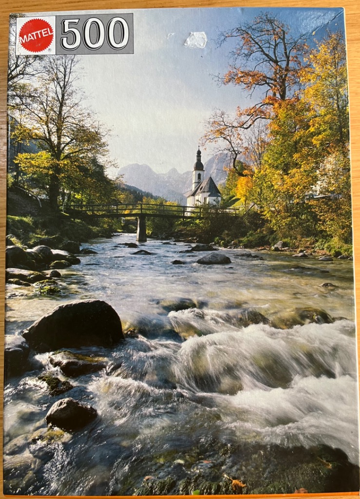 Scenic autumn landscape featuring a rushing river, a footbridge, and a church with mountain peaks in the background.