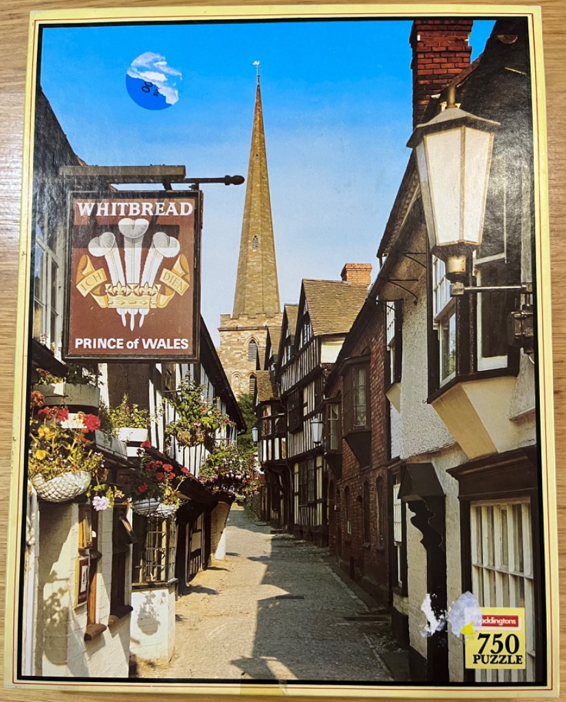 A narrow English street with half-timbered buildings and a pub sign leading toward a church spire under a blue sky.