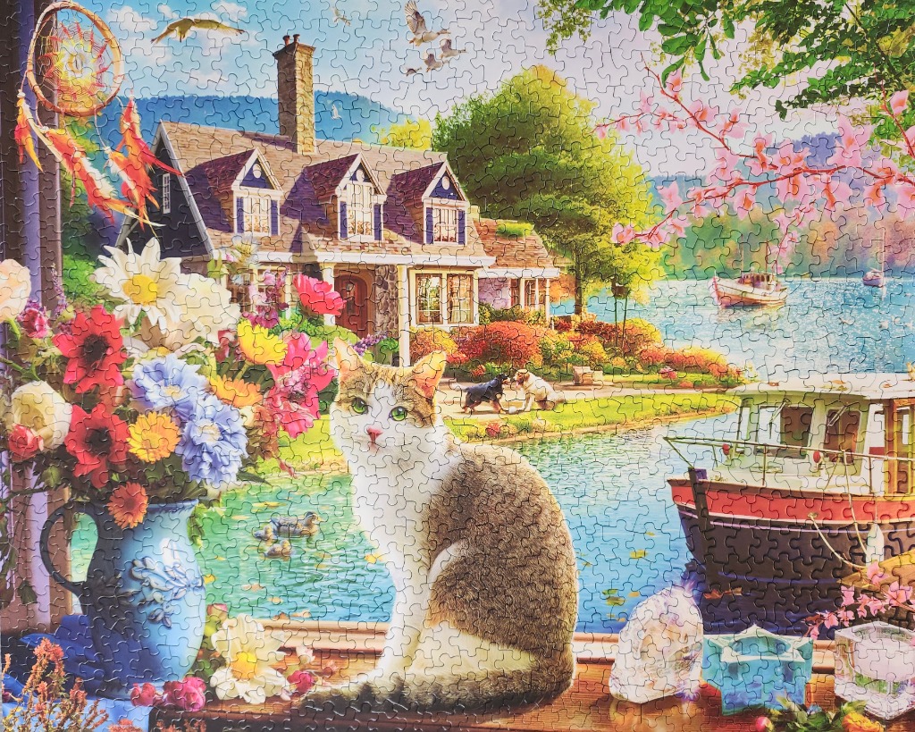 A tabby cat sits on a windowsill overlooking a lake house with a boat, flowers, and a dreamcatcher.