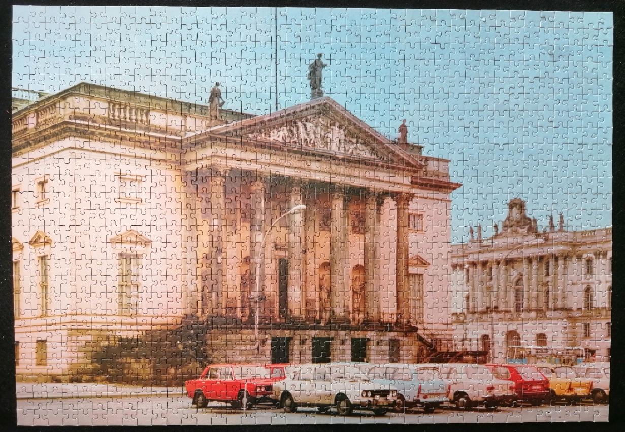 A street-level view of a classical European building with columns and sculptures, flanked by vintage cars in the foreground.