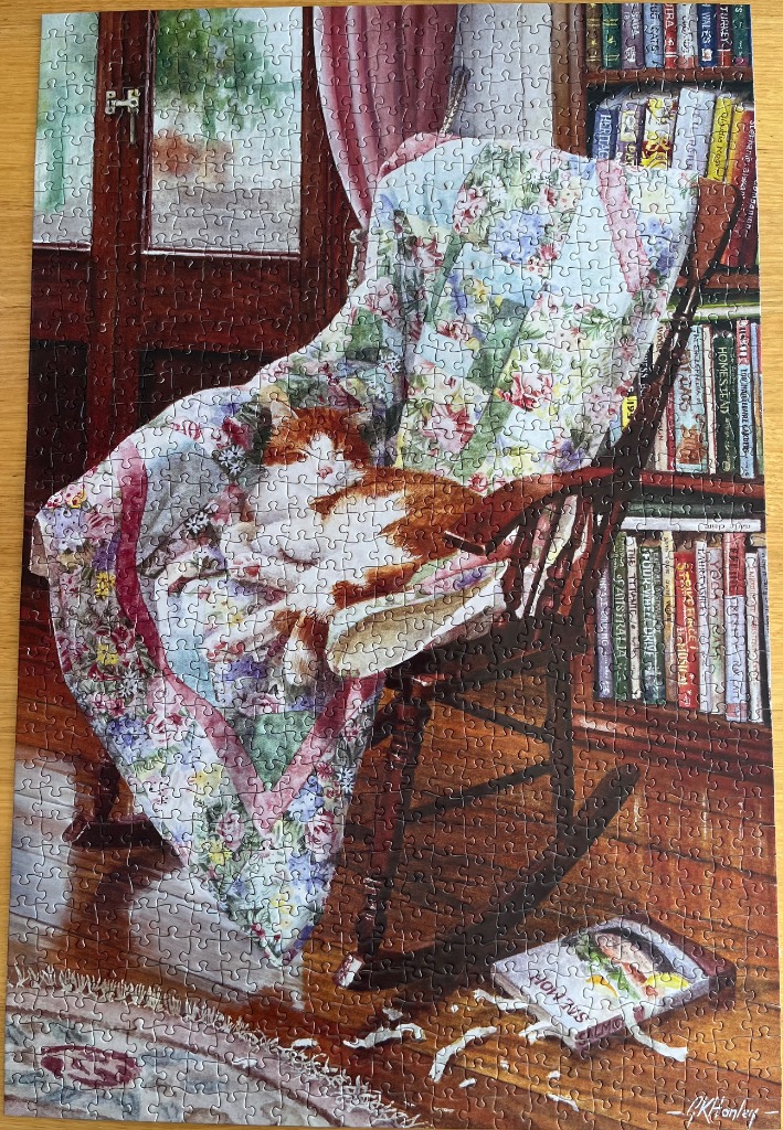 A ginger and white cat sleeping on a quilt in a wooden rocking chair next to a bookshelf.