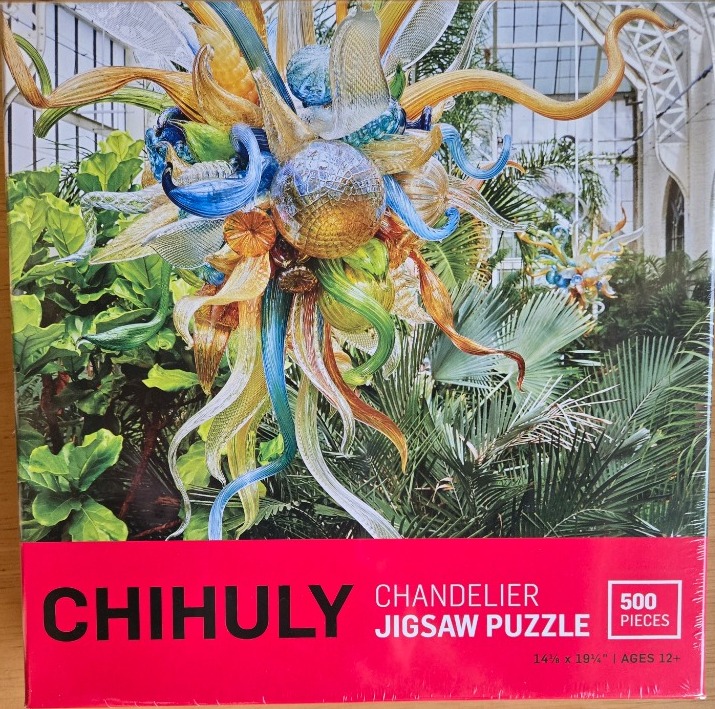 A vibrant, multi-colored glass sculpture hangs in a lush greenhouse setting.