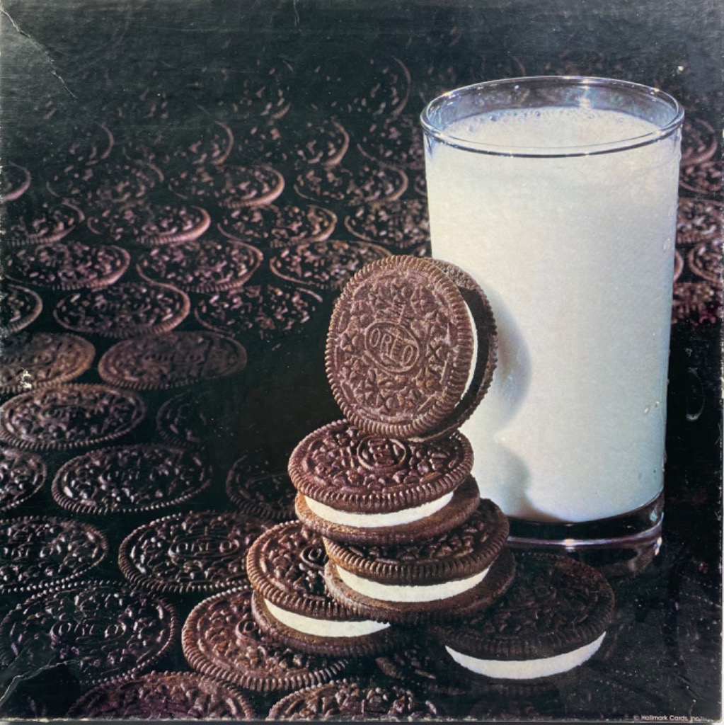 A stack of chocolate sandwich cookies beside a tall glass of milk, set against a background of scattered cookies.