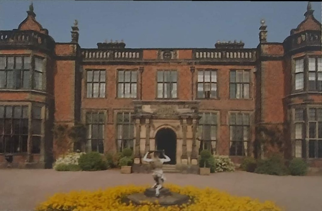 A large, symmetrical red-brick Jacobean-style manor house with a front garden statue and flowerbed.
