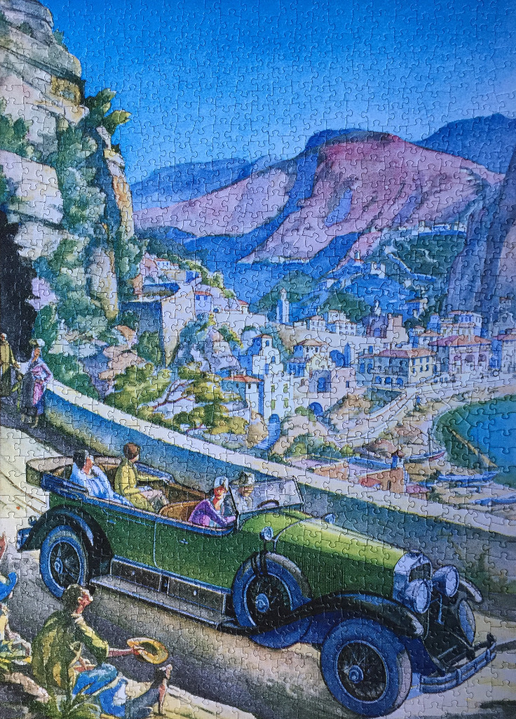 Vintage green touring car driving along a coastal cliffside road overlooking a seaside town.