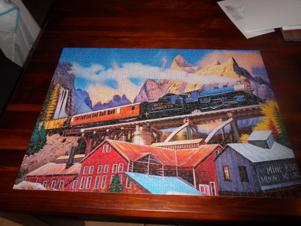 A steam train crosses a high wooden trestle bridge above a mining facility set against rugged mountain peaks.