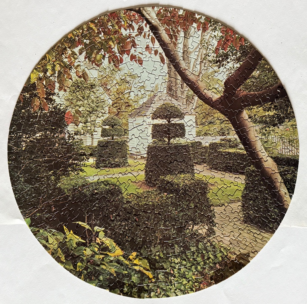 Circular photograph of a manicured garden with topiary shrubs and a white house in the background.