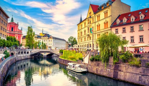 A sunny riverside view of Ljubljana, Slovenia, featuring the Triple Bridge and colorful historic buildings.