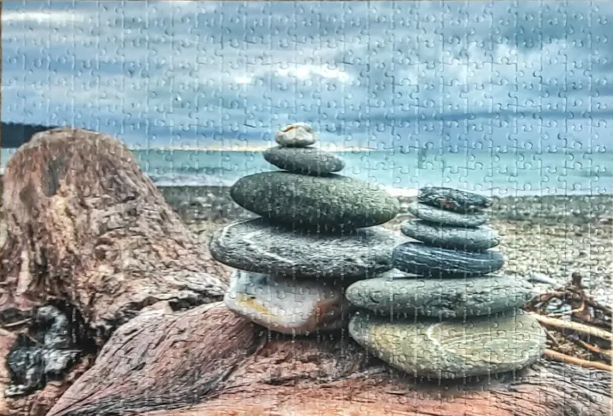 Two stone cairns balanced on a piece of driftwood on a beach under a cloudy sky.