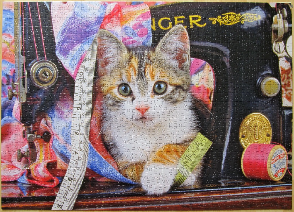 A calico kitten peering from inside an antique black sewing machine, surrounded by colorful fabric and notions.