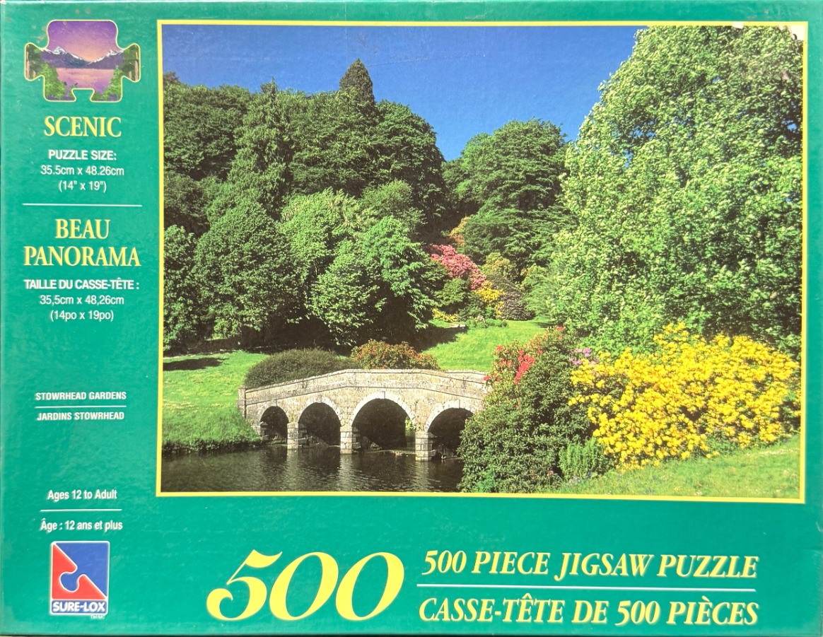 A stone bridge arches over a calm pond in a lush garden filled with green trees and yellow and pink flowering shrubs.
