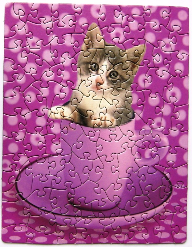 A small kitten peeking out from inside a purple teacup against a purple polka-dot background.