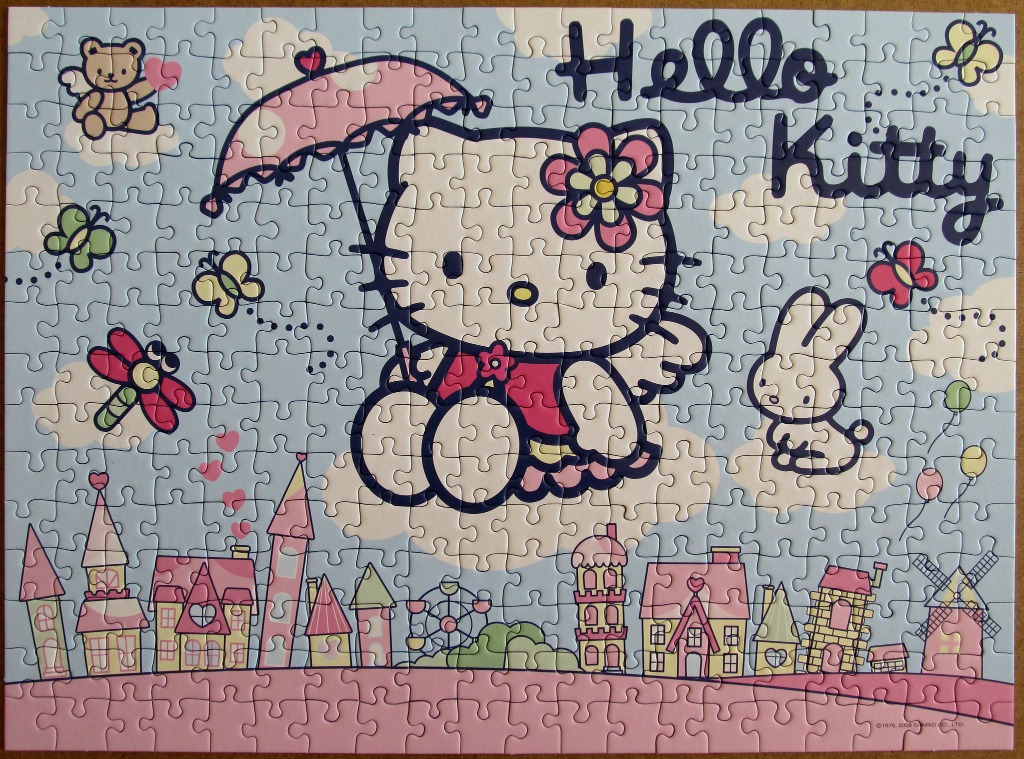 Hello Kitty character flying with a pink umbrella over a whimsical village