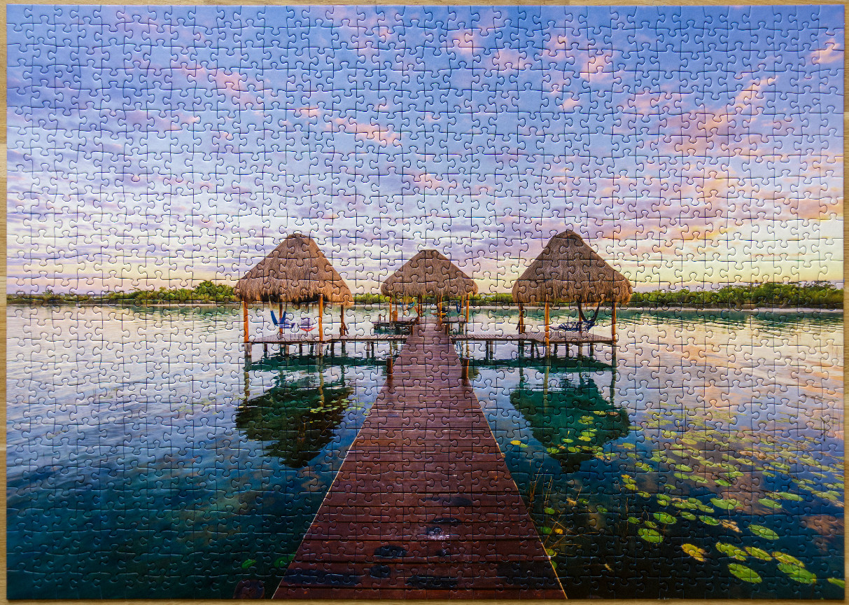 A wooden dock leads toward thatched-roof gazebos over a calm, clear lake at sunrise.