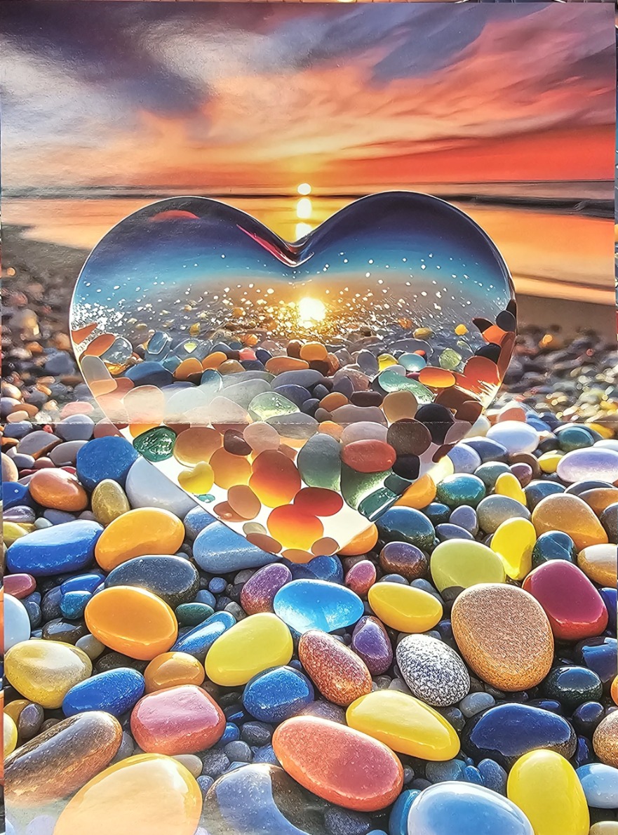 A heart-shaped glass object sitting on a beach of colorful polished stones at sunset.