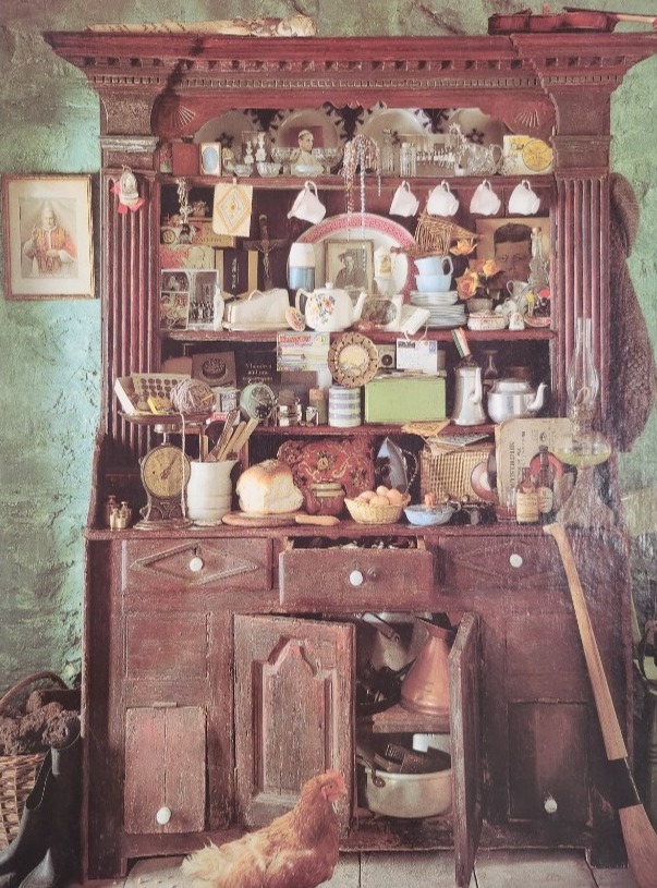 A rustic wooden hutch overflowing with eclectic household objects, curios, and a chicken standing on the floor in front.