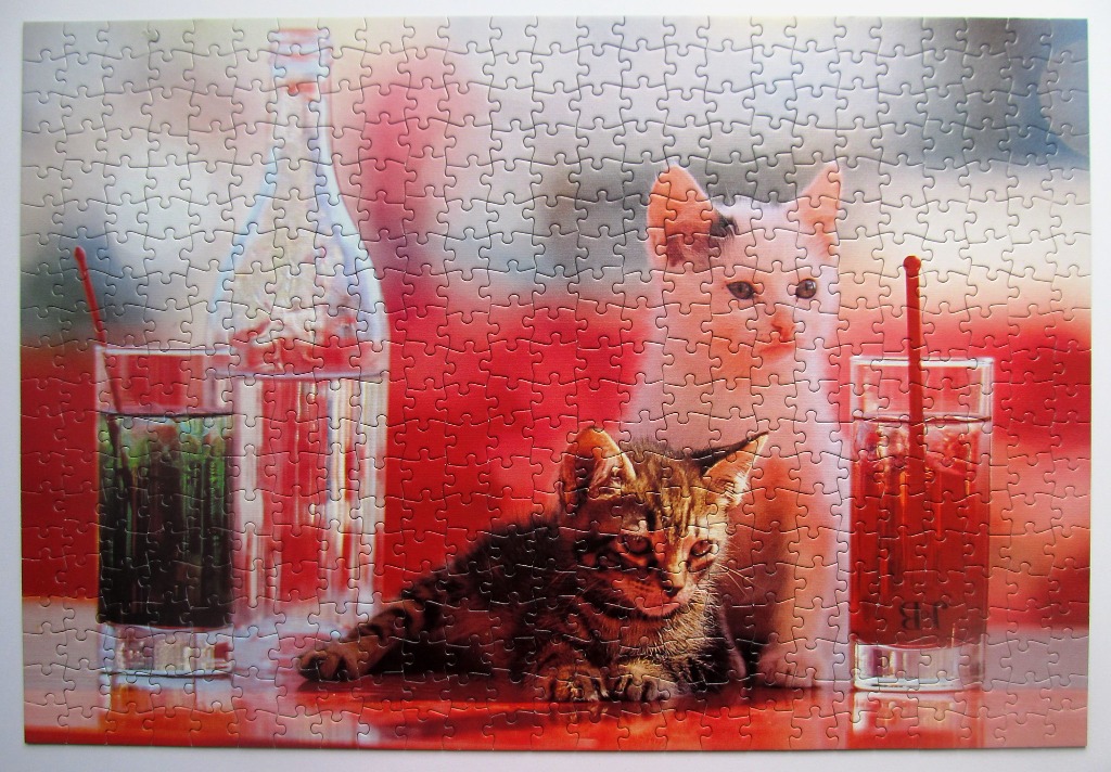 A white kitten and a tabby kitten sitting next to a glass bottle and two glasses with red liquid.