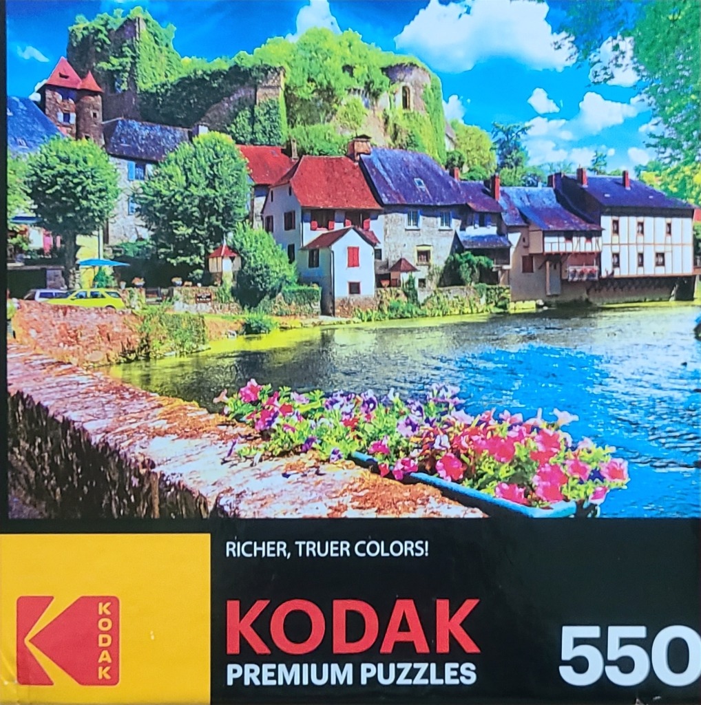 A sunny European village by a river with stone houses, a hillside castle, and pink flowers in the foreground.