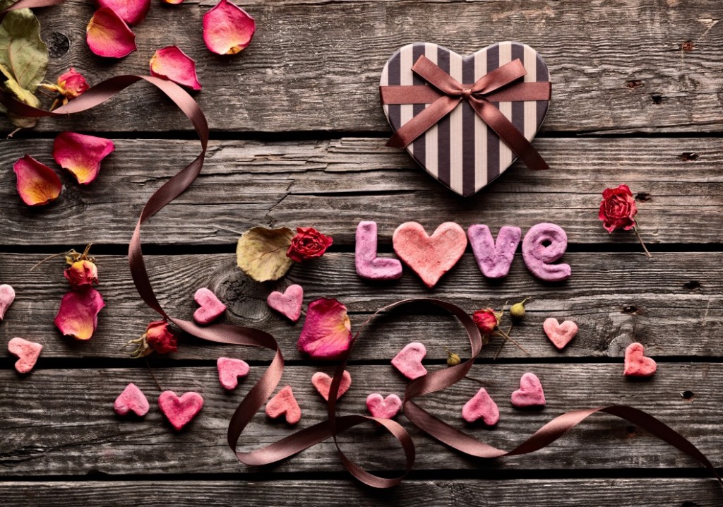 A heart-shaped gift box, decorative hearts, rose petals, and a ribbon on a rustic wooden surface.