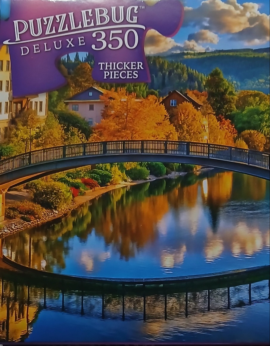 A peaceful autumnal scene featuring a dark arched footbridge spanning a calm lake with reflections of trees and buildings.