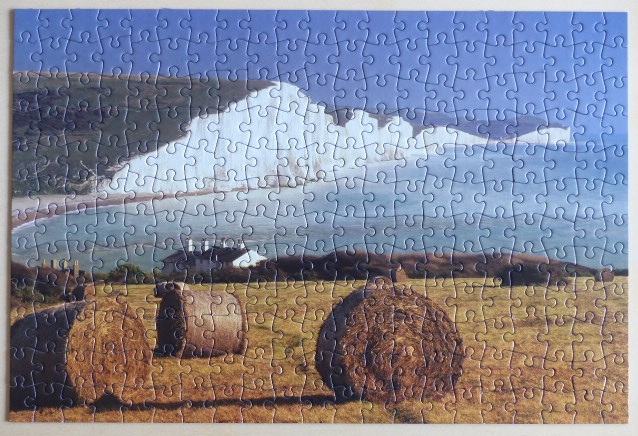 A sunny, coastal landscape featuring white chalk cliffs above the sea and round hay bales in a golden field.