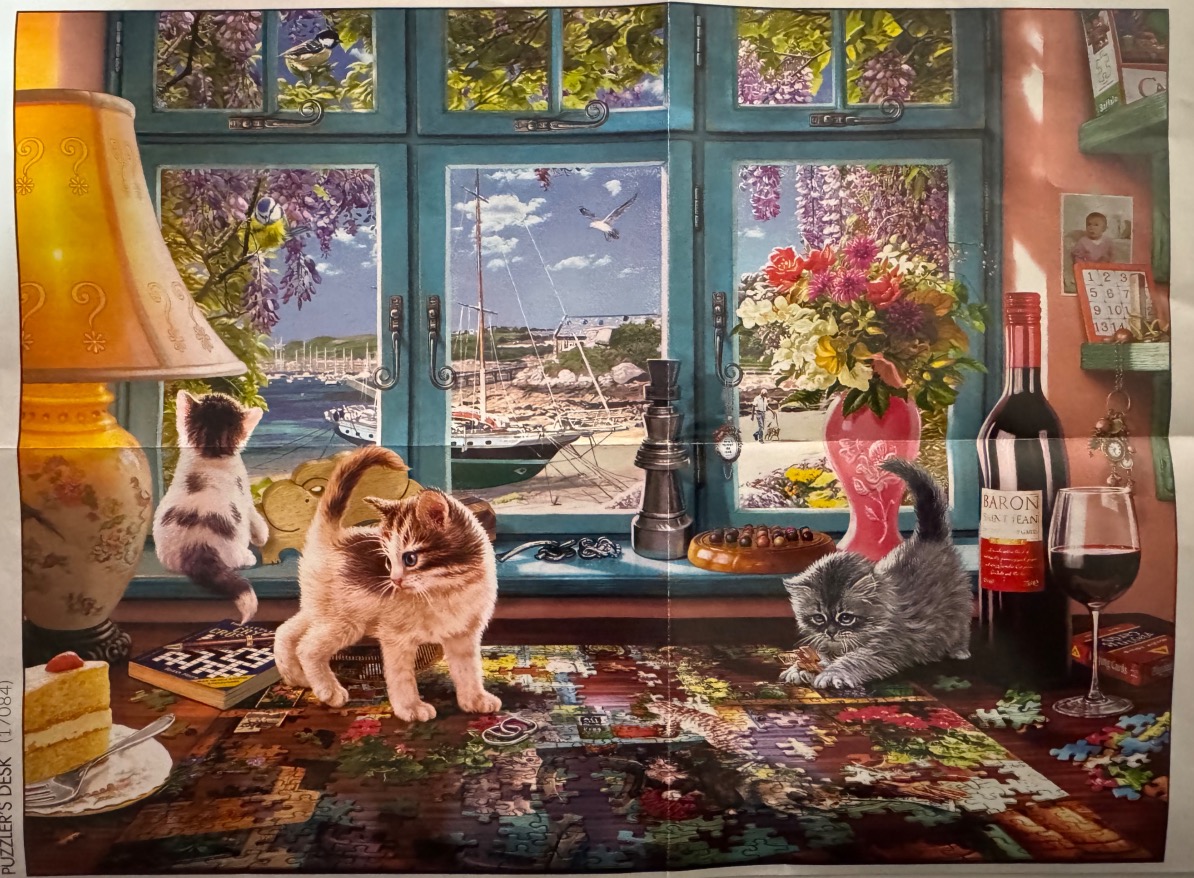 Three kittens playing on a table with a jigsaw puzzle in front of a window overlooking a seaside harbor.