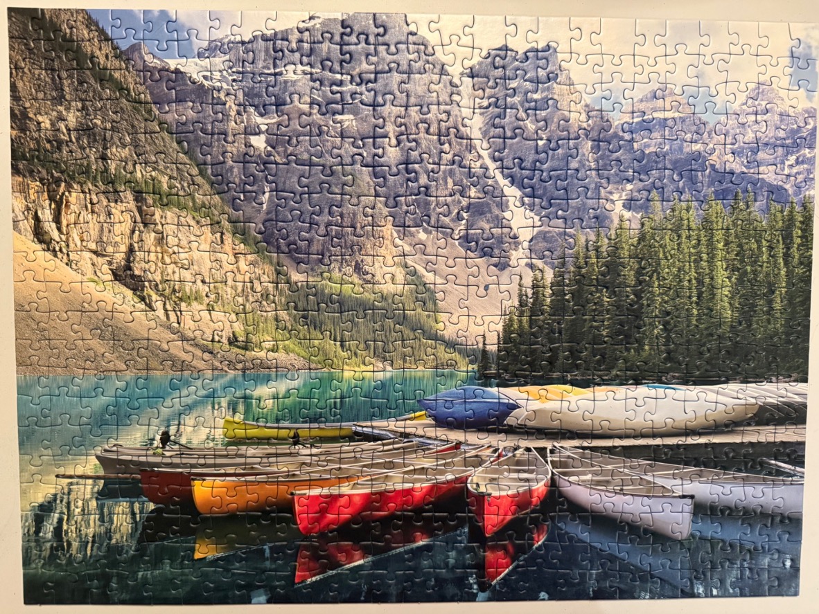 A row of colorful canoes docked at a lake beneath tall, jagged mountain peaks and evergreen trees.