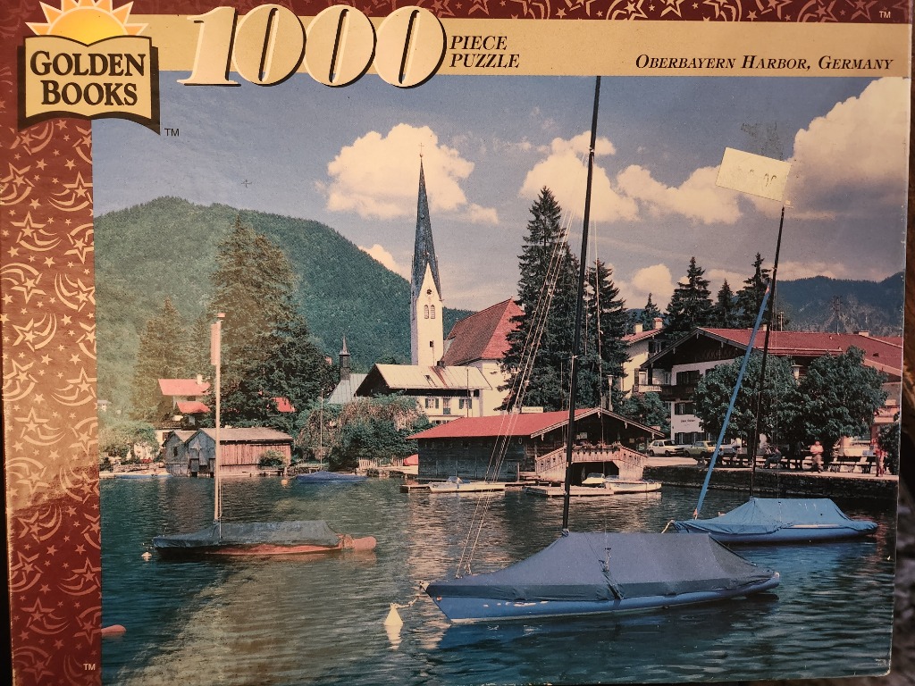 A photograph of a harbor in Oberbayern, Germany, featuring sailboats, a historic church, and mountainous scenery.