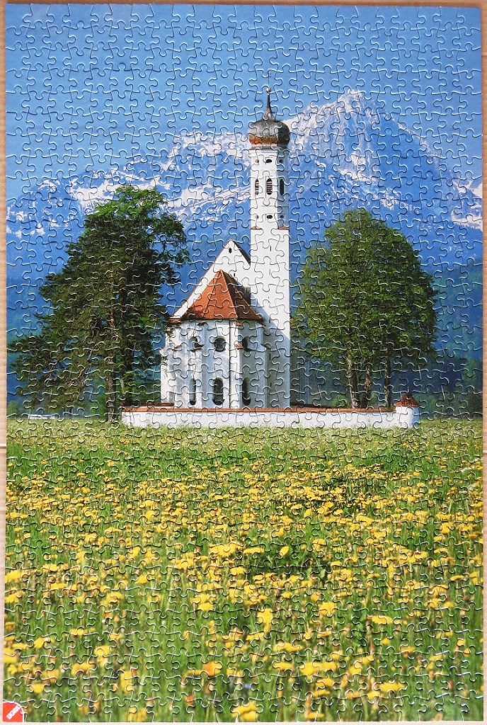 A white church with a tall steeple stands in a meadow of yellow flowers against a backdrop of snow-capped mountains.