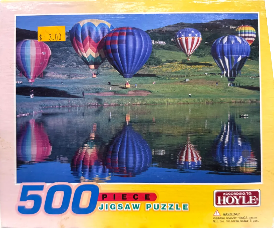 Multiple colorful hot air balloons floating over a grassy field and a reflective lake.
