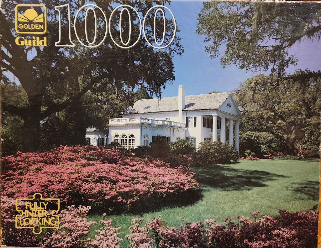 A stately white colonial-style mansion surrounded by blooming pink azaleas and large oak trees with Spanish moss.