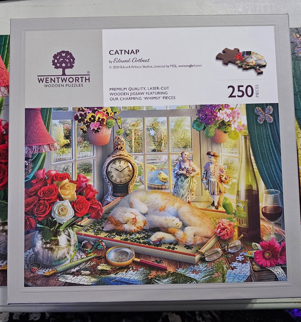 A ginger cat sleeping on a jigsaw puzzle board in a sunny window-filled room with flowers and figurines.