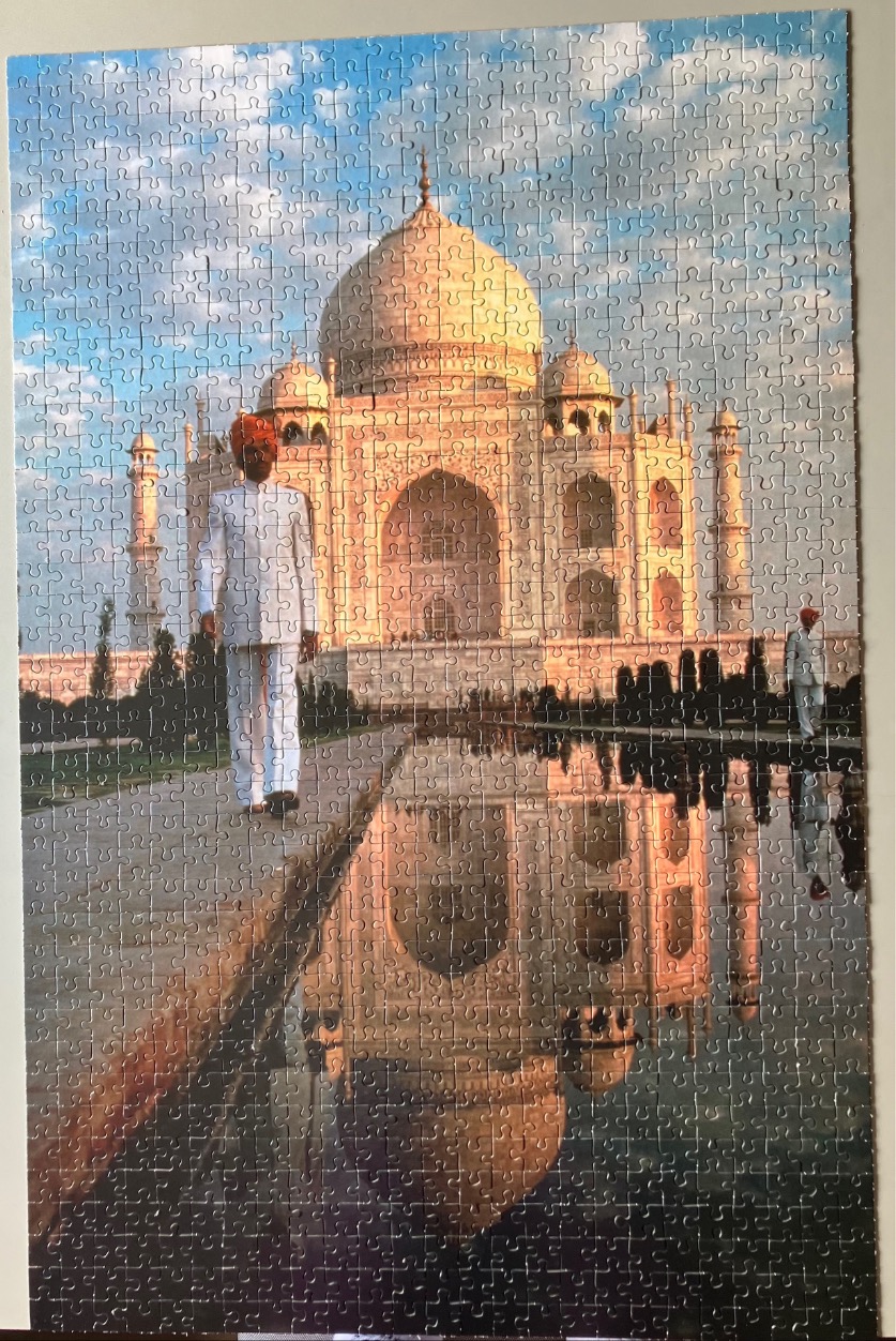 A man in traditional white attire and a red turban walks past the Taj Mahal, which is reflected in a calm pool.