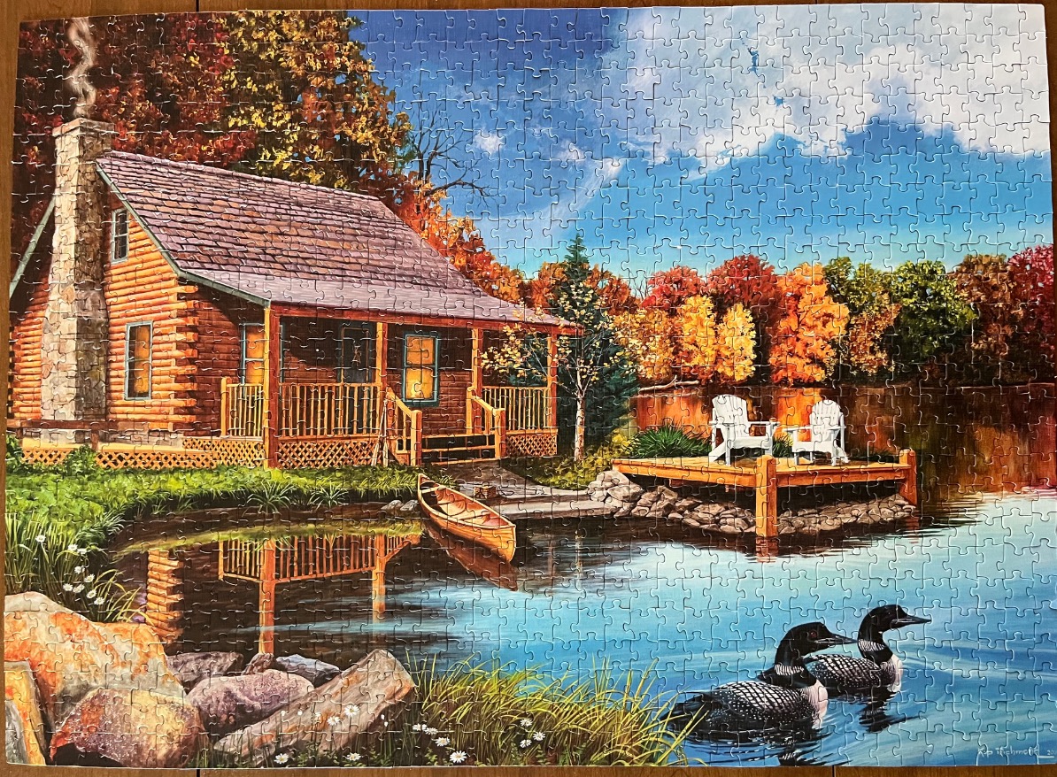 A log cabin sits beside a lake with a canoe, a wooden deck, and two loons in the water during autumn.